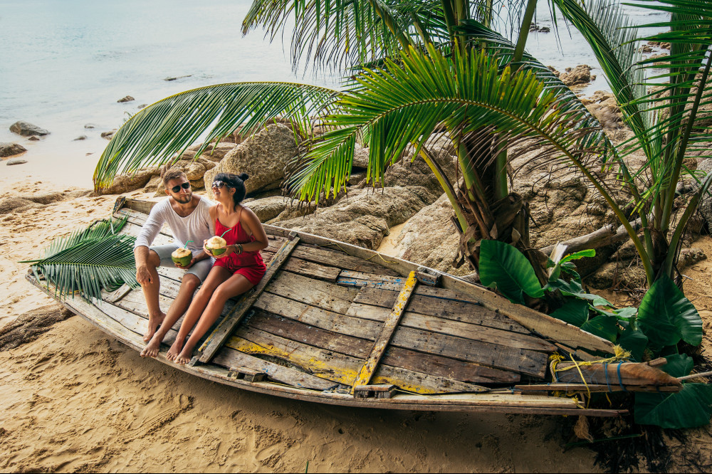 A couple relaxing in a old pirogue (boat) tilted on the beach under some palm trees.