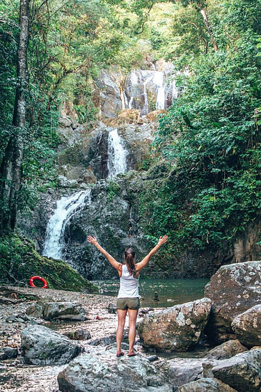 A woman with arms outstretched looking at waterfall and pools in which people are swimming