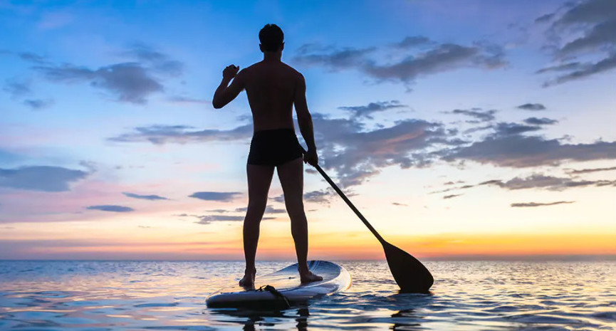 A man with his back turned paddle boarding at sunset