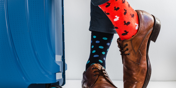 A standing blue suitcase next to a person wearing one multicoloured red and blue sock each in brown laced shoes