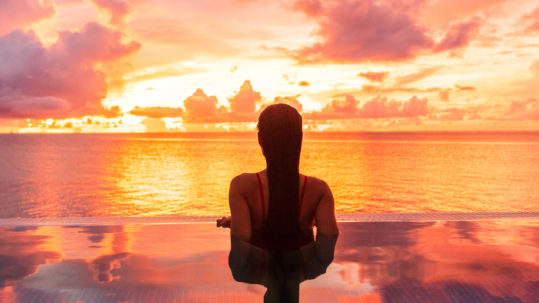 Young woman at infinity pool edge looking at sunset