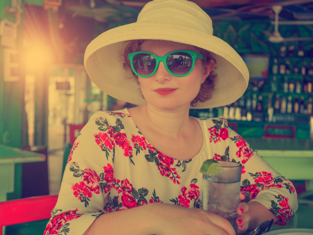 A young white woman in floral blouse, shades and white hat holding a glass of water with straw and lemon