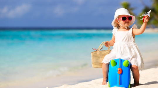 Little girl dressed in white summer dress sitting on a suitcase on a beach holding a miniature plane