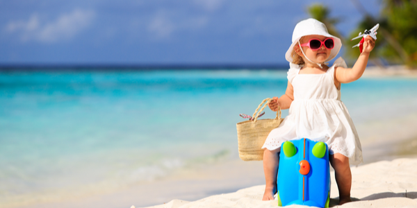 Little girl dressed in white summer dress sitting on a suitcase on a beach holding a miniature plane